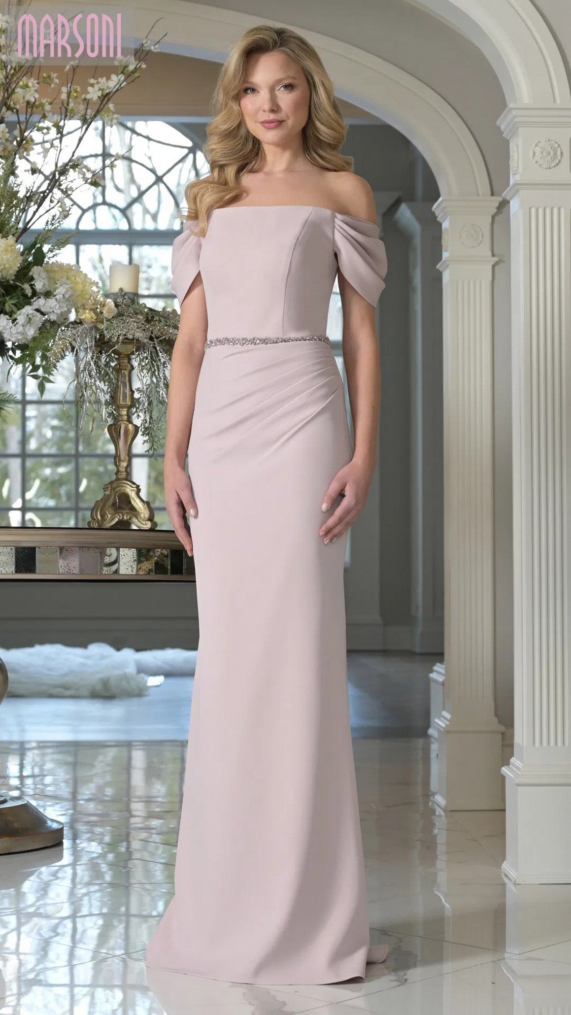 A woman stands indoors wearing the Marsoni MV1352 long formal dress in mauve, featuring an off-shoulder design and beaded waistband. Ornate columns and floral decor create an elegant backdrop.